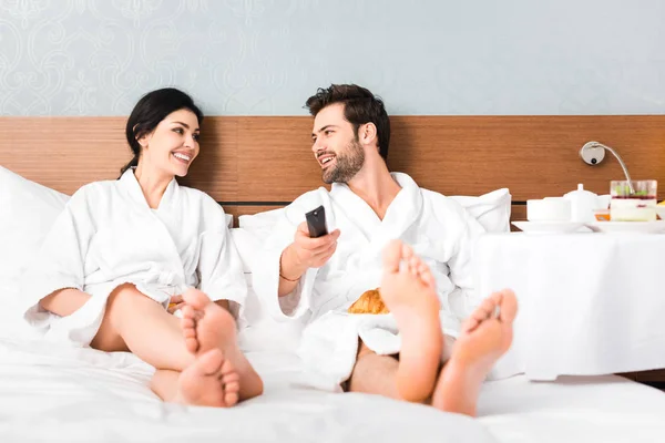 Selective focus of cheerful man holding remote controller while looking at woman in hotel room — Stock Photo