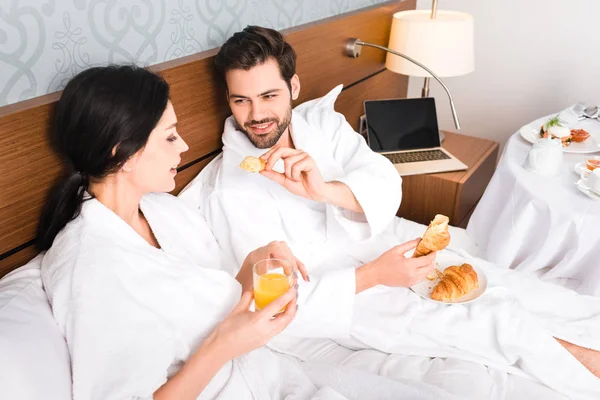 Allegro uomo che tiene croissant mentre guarda donna attraente con vetro di succo d'arancia — Foto stock