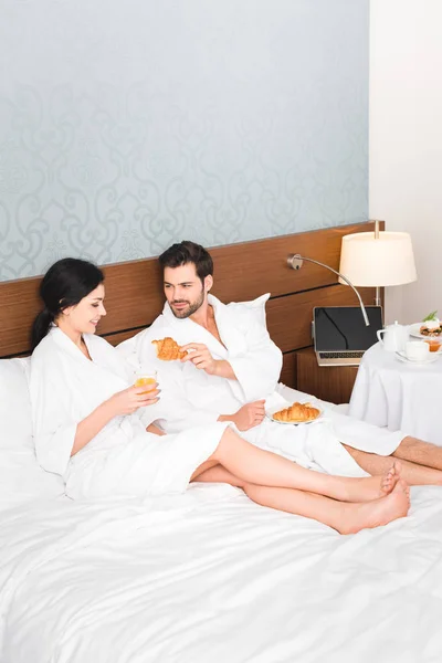 Barbudo hombre celebración croissant cerca feliz mujer con vaso de jugo de naranja - foto de stock