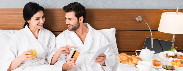 Foto panorámica de la mujer feliz señalando con el dedo en el desayuno cerca de hombre con tarjeta de crédito y tableta digital - foto de stock