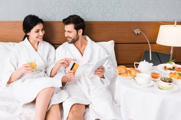 Mujer feliz señalando con el dedo en el desayuno cerca de hombre con tarjeta de crédito y tableta digital - foto de stock