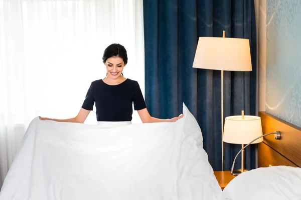 Cheerful housemaid holding white bed sheet in hotel room — Stock Photo