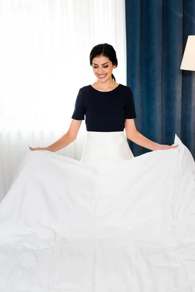 Happy housemaid holding white bed sheet in hotel room — Stock Photo