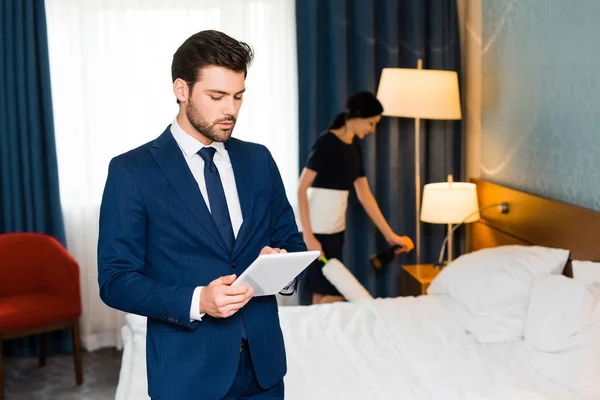 Enfoque selectivo de hombre guapo utilizando tableta digital cerca de criada en la habitación de hotel - foto de stock