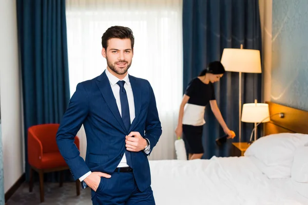 Heureux réceptionniste debout avec la main dans la poche près de la bonne dans la chambre d'hôtel — Photo de stock