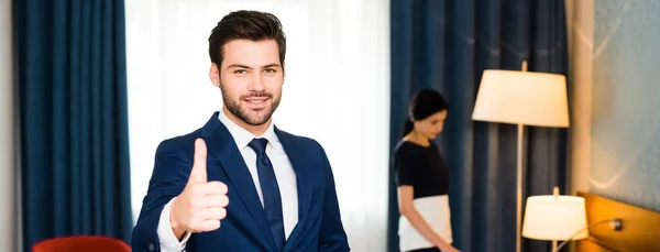 Foto panorámica de la recepcionista feliz mostrando el pulgar hacia arriba cerca de la criada en la habitación del hotel - foto de stock