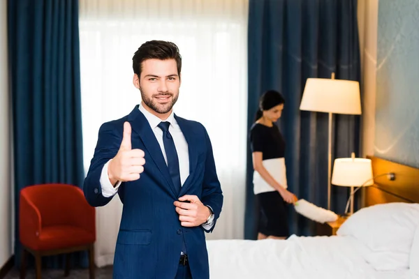 Enfoque selectivo de la recepcionista feliz mostrando el pulgar hacia arriba cerca de la criada en la habitación del hotel - foto de stock