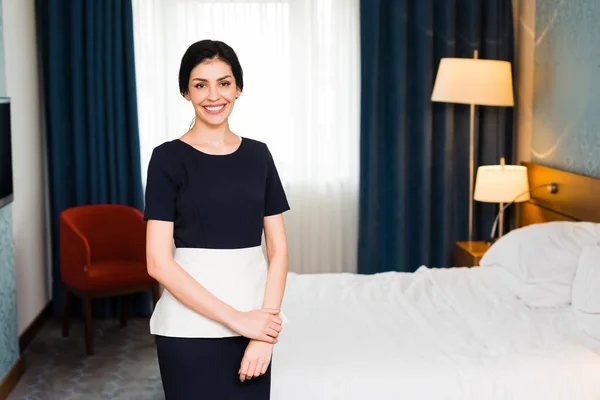 Empregada doméstica alegre sorrindo enquanto olha para a câmera no quarto do hotel — Fotografia de Stock