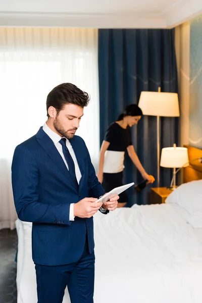 Schöner Mann im Anzug mit digitalem Tablet nahe Dienstmädchen im Hotelzimmer — Stockfoto