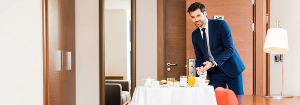 Panoramic shot of cheerful receptionist in formal wear gesturing near breakfast in hotel room — Stock Photo