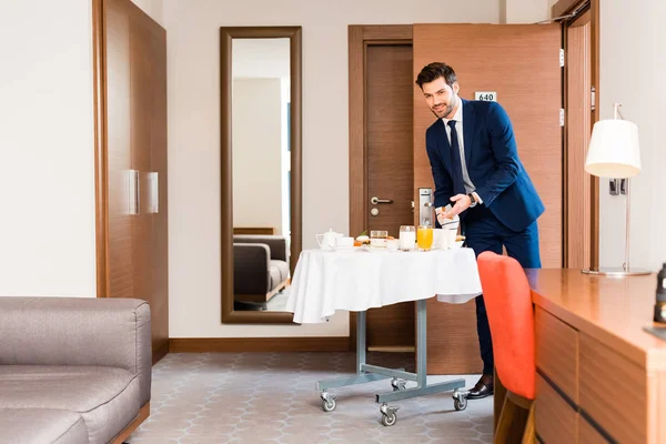 Cheerful receptionist in formal wear gesturing near breakfast in hotel room — Stock Photo
