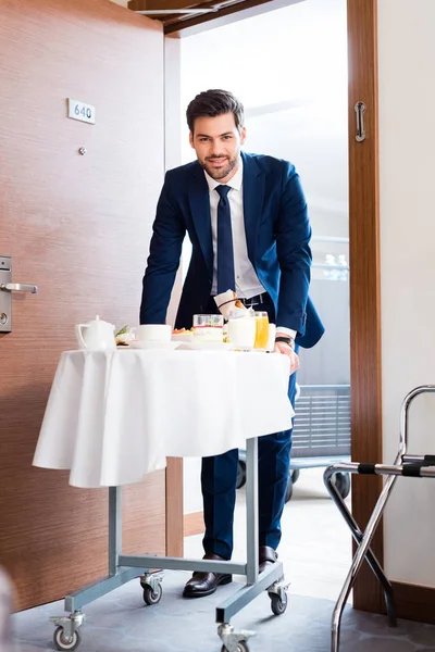 Selektiver Fokus der fröhlichen Empfangsdame, die das Frühstück auf dem Hotelwagen bringt — Stockfoto