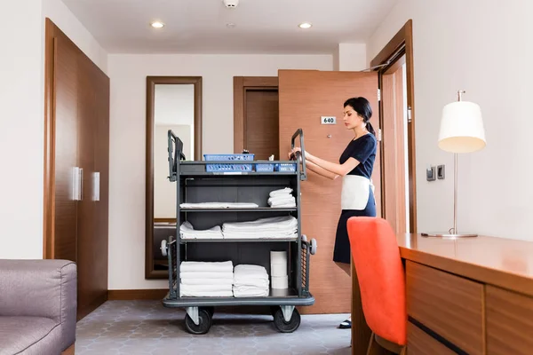 Attractive brunette housemaid in uniform standing near cleaning trolley with white towels — Stock Photo