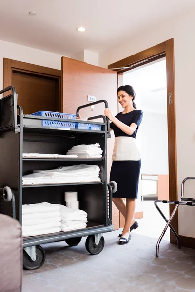 Low angle view of happy housemaid in uniform standing near cleaning trolley with white towels — Stock Photo