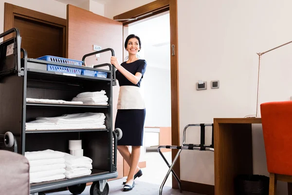 Visão de baixo ângulo da empregada doméstica alegre em pé uniforme perto do carrinho de limpeza com toalhas brancas — Fotografia de Stock