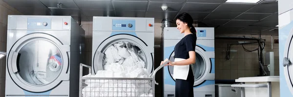 Panoramic shot of happy maid looking at dirty bedding while standing near metallic cart — Stock Photo