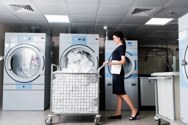 Beautiful and happy maid looking at dirty bedding while standing near metallic cart — Stock Photo