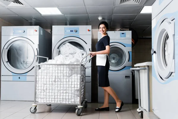 Cheerful maid standing near metallic cart with bedding in laundry — Stock Photo