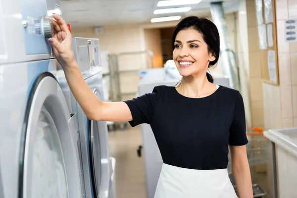 Selective focus of happy maid touching button on washing machine in laundry — Stock Photo