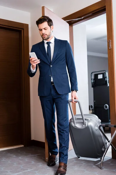 Handsome businessman looking at smartphone while standing with suitcase in hotel — Stock Photo