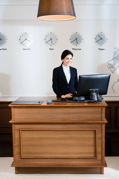 Fröhliche brünette Empfangsdame in formeller Kleidung, die auf den Computermonitor im Hotel blickt — Stockfoto