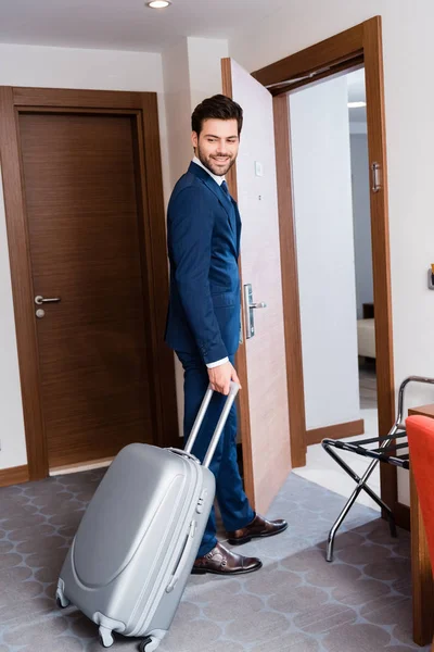 Alegre hombre de negocios en traje sonriendo mientras está de pie con la maleta en la habitación de hotel - foto de stock