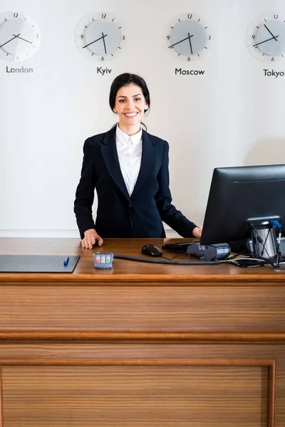 Fröhliche brünette Empfangsdame in formeller Kleidung, die in der Nähe des Computermonitors im Hotel steht — Stockfoto