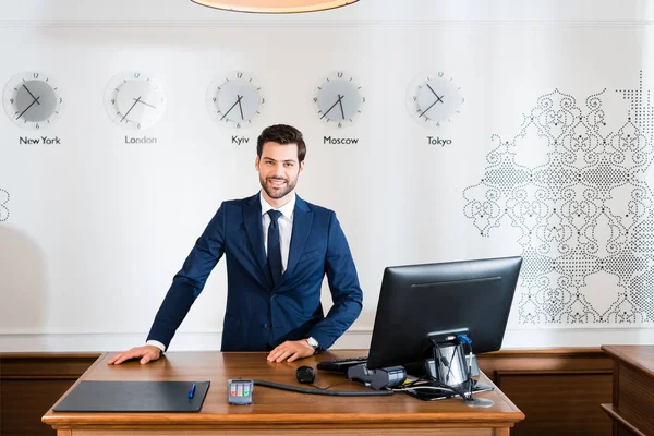 Recepcionista alegre no terno que está perto do monitor de computador no hotel — Fotografia de Stock