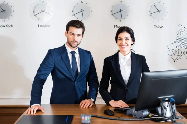 Cheerful woman near serious receptionist in suit standing at reception desk — Stock Photo