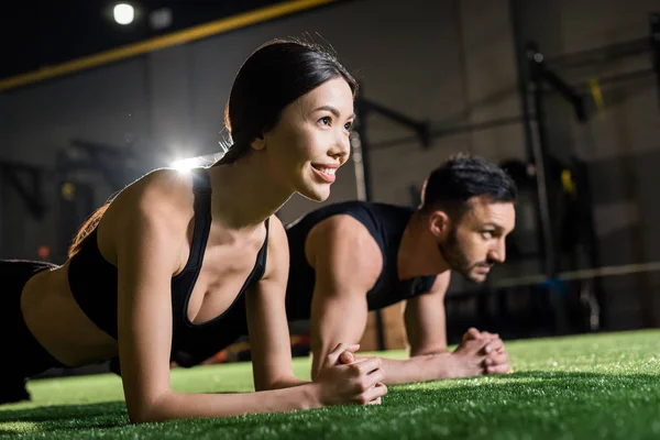 Selective focus of cheerful woman smiling while doing plank exercise near man on grass — Stock Photo