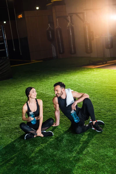 Glückliche Frau, die mit einem hübschen Mann auf Gras sitzt und eine Sportflasche mit Wasser in der Hand hält — Stockfoto