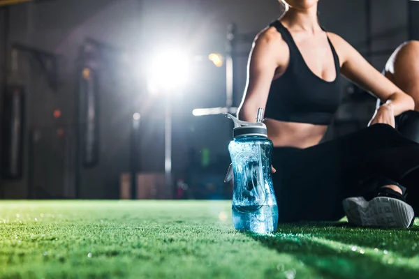 Abgeschnittene Ansicht einer athletischen Frau, die auf Gras sitzt und eine Sportflasche in der Hand hält — Stockfoto
