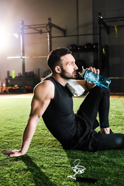 Hombre atlético guapo beber agua mientras está sentado en la hierba cerca de teléfonos inteligentes y auriculares - foto de stock