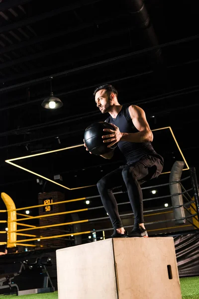 Low angle view of serious man doing squat on squat box while holding ball — Stock Photo