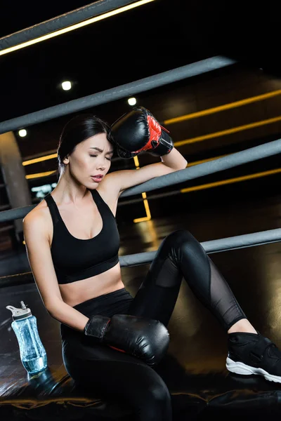 Woman in boxing gloves touching head near sport bottle in gym — Stock Photo