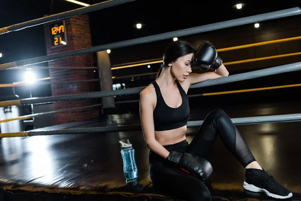 Tired woman in boxing gloves touching head near sport bottle in gym — Stock Photo