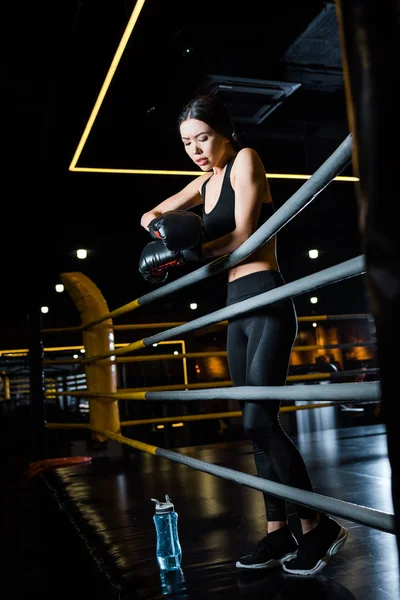 Low angle view of athletic woman looking at boxing gloves while standing near sport bottle — Stock Photo