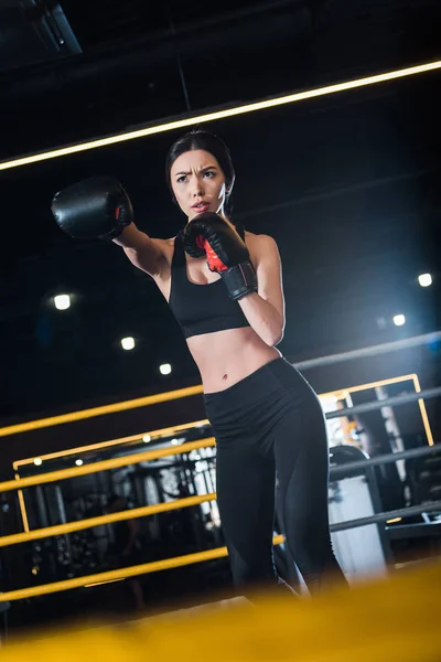 Selective focus of young woman boxing while standing in boxing gloves in gym — Stock Photo