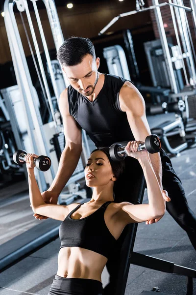 Enfoque selectivo de la mujer atlética joven haciendo ejercicio con pesas cerca de entrenador guapo en el gimnasio - foto de stock