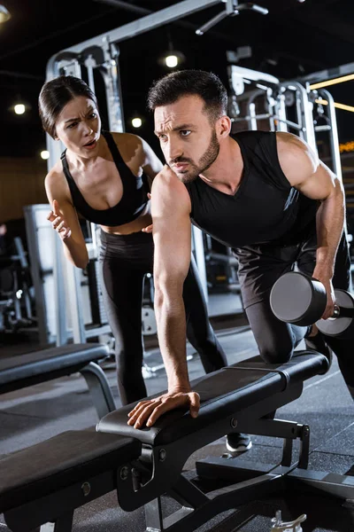 Vista de ángulo bajo de entrenador atractivo de pie cerca de hombre guapo haciendo ejercicio con mancuerna - foto de stock