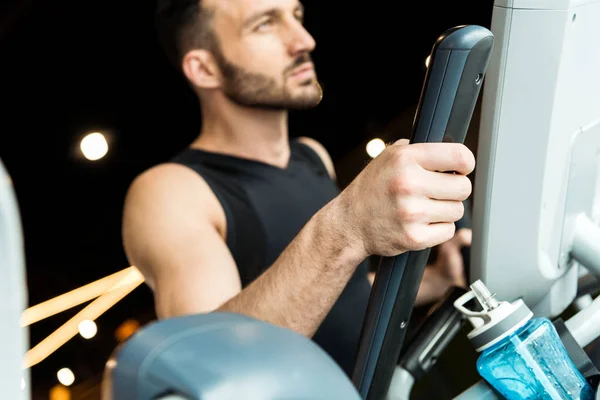 Foco seletivo de garrafa de esporte perto de homem atlético bonito trabalhando em bicicleta de exercício — Fotografia de Stock