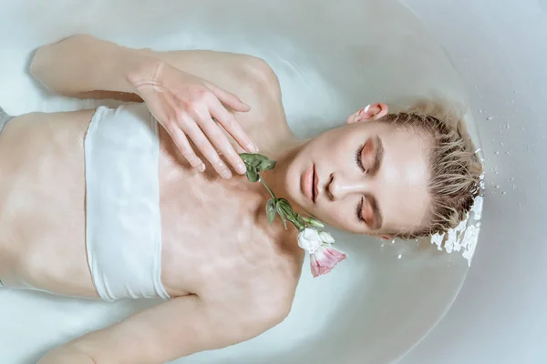 Vista superior da mulher macia com os olhos fechados suavemente segurando flor em água clara na banheira — Fotografia de Stock
