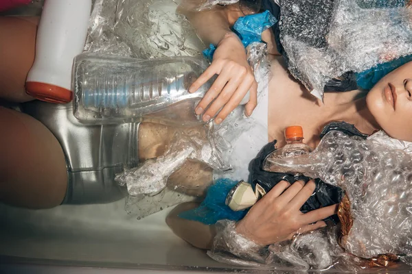 Обрезанный вид молодой женщины в серебряном белье позирует в ванне с пластиковыми отходами, эко-концепция — стоковое фото