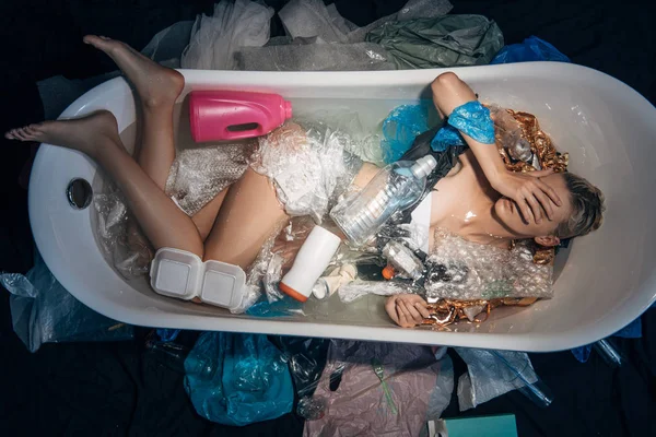 Vista superior de la mujer joven acostada en la bañera con facepalm entre la basura, concepto de contaminación ambiental - foto de stock