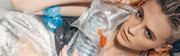 Tiro panorámico de mujer húmeda triste sosteniendo peces de colores en bolsa de plástico entre la basura en la bañera - foto de stock