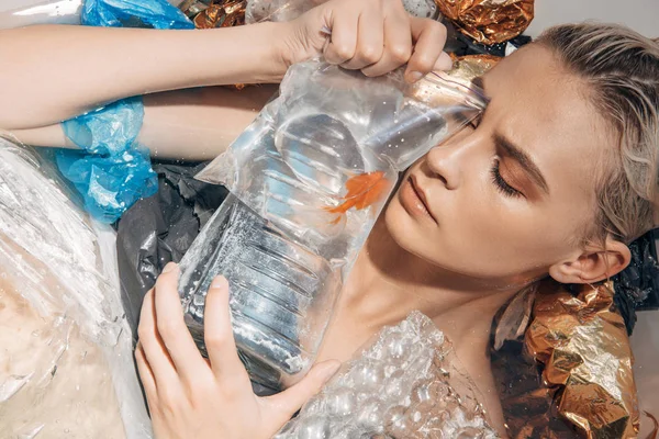 Сумна мокра жінка з закритими очима тримає золоту рибу в поліетиленовому пакеті серед сміття у ванні — стокове фото