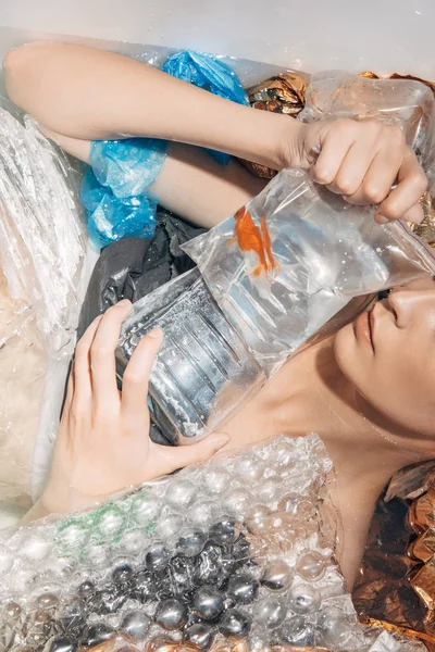 Vista recortada de la mujer mojada sosteniendo peces de colores en bolsa de plástico entre la basura en la bañera - foto de stock