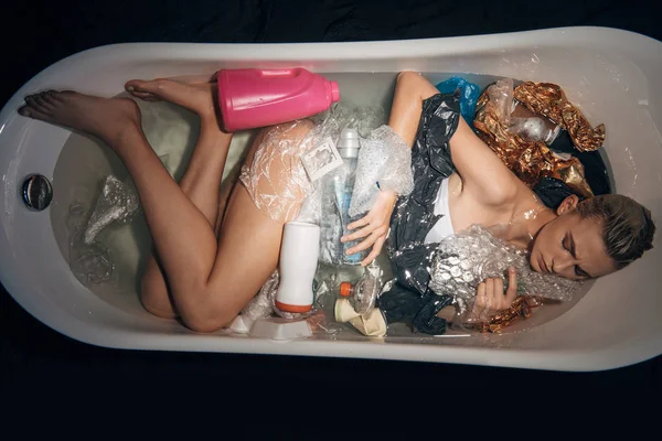 Top view of young sad woman lying in bathtub with trash isolated on black, environmental pollution concept — Stock Photo