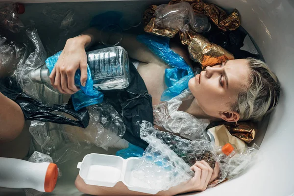 Top view of young sad woman lying in bathtub with trash, environmental pollution concept — Stock Photo