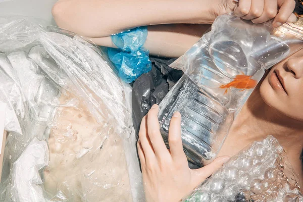 Vista cortada de jovem segurando peixinho dourado na banheira com lixo plástico, conceito de poluição ambiental — Fotografia de Stock